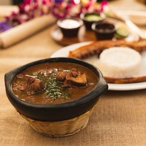 Hearty bean stew with meat in a black bowl, with rice and plantains in the background.