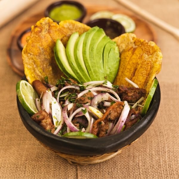 A bowl of fried plantains, sliced avocado, meat, and red onions with lime wedges.