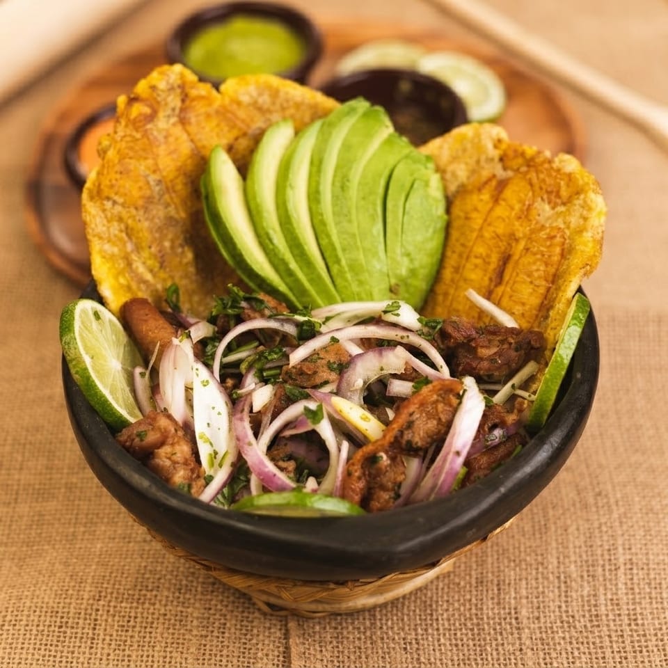 A bowl of fried plantains, sliced avocado, meat, and red onions with lime wedges.
