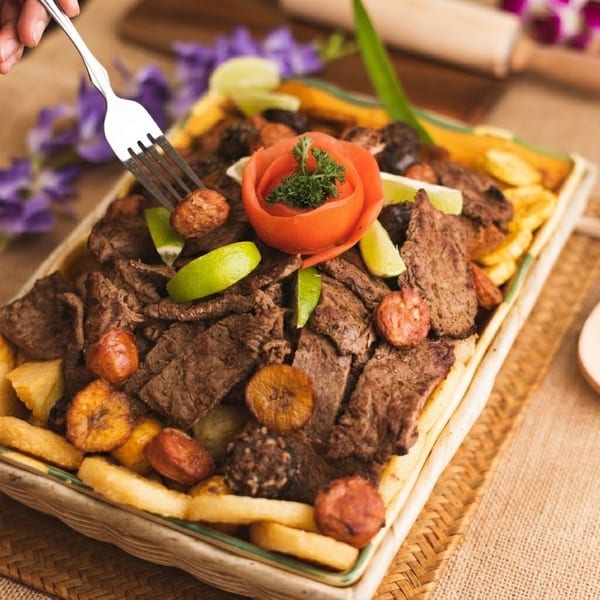 A fork lifts a piece of sausage from a platter of sliced meat, plantains, and fries.