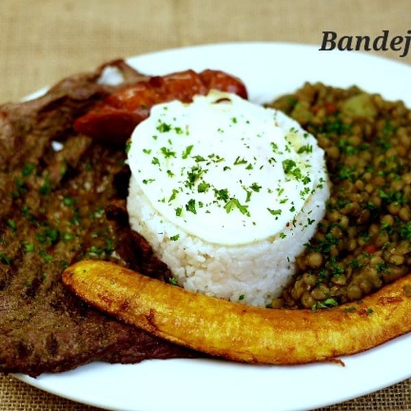 Bandeja valluna with grilled steak, rice, fried egg, plantain, and lentils on a white plate.