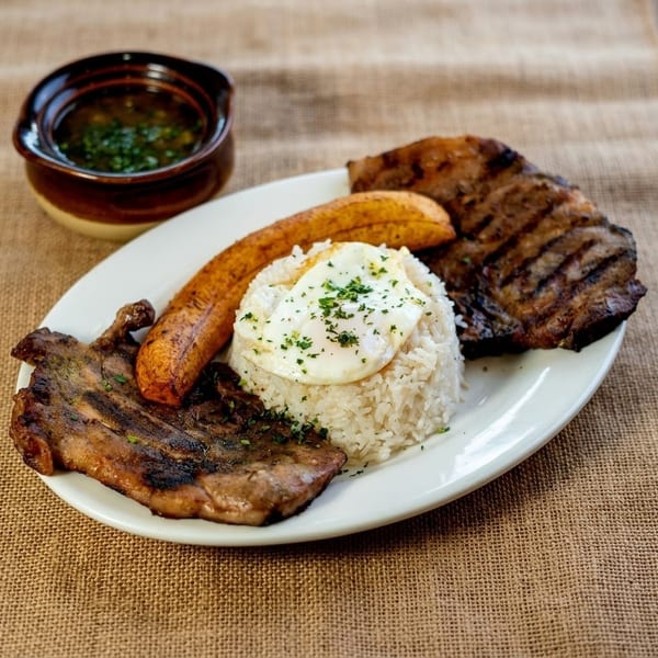 Grilled steak, rice with a fried egg, plantains, and a side of soup on a white plate.